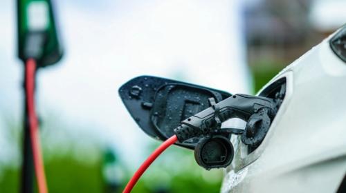 En elbil ansluten till en laddstation med en orange kabel under en molnig himmel.