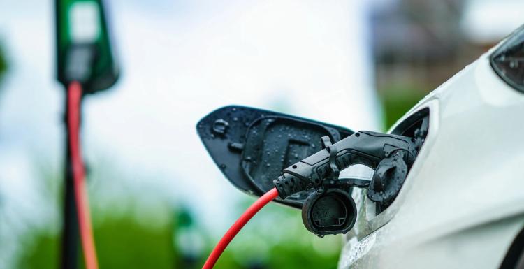 Sähköauto yhdistettynä latausasemaan oranssilla kaapelilla pilvisen taivaan alla.