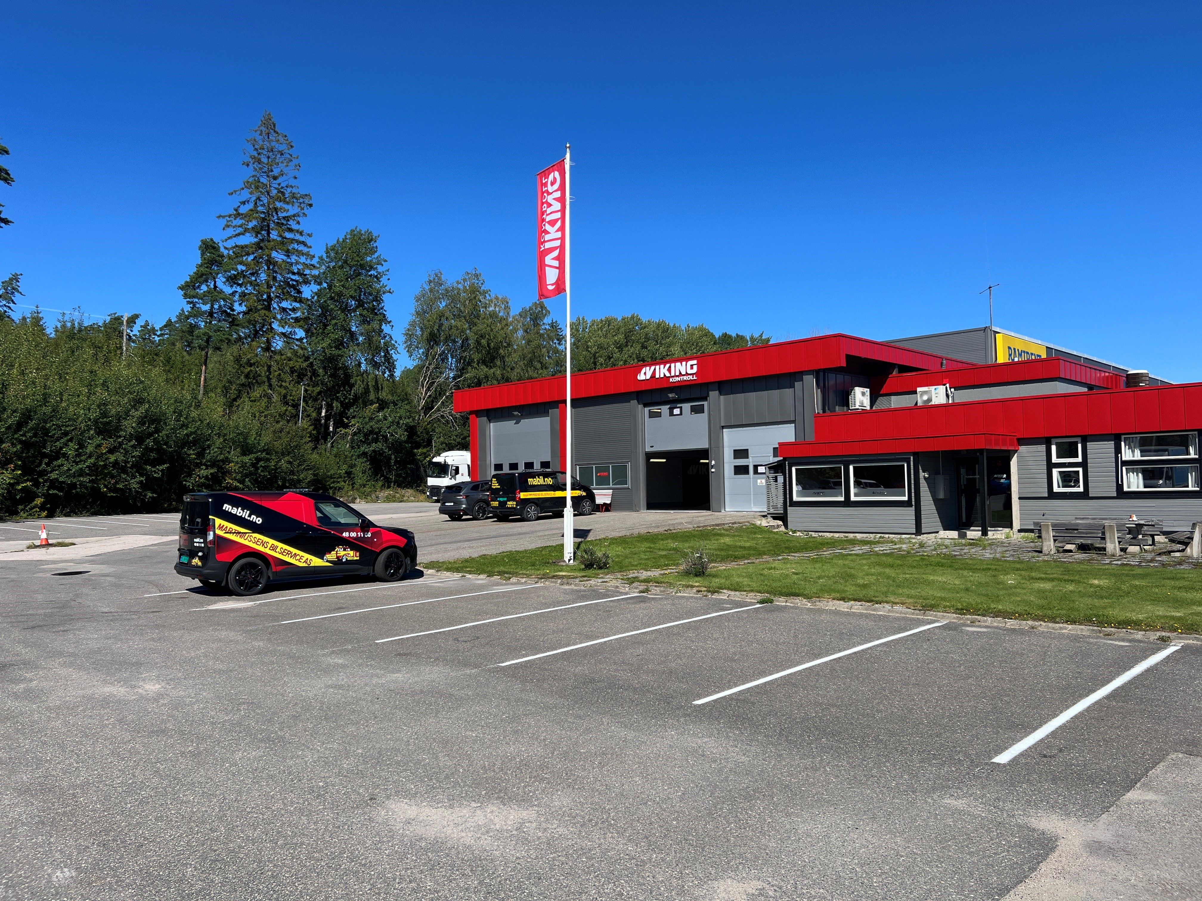 Et bilserviceanlegg med en rød og grå bygning, en merkevarebil, et flagg og en tom parkeringsplass på en solrik dag. Skogområde i bakgrunnen.