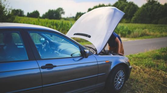 Henkilö tutkimassa auton moottoritilaa konepelti auki maaseututien laidassa.