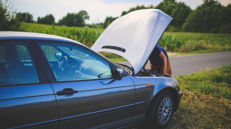 A person examining the engine compartment of a car with the bonnet open on the side of a rural road.