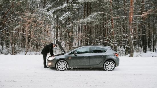 Person som undersøker motoren til en grå bil som ikke starter på en snødekt vei.