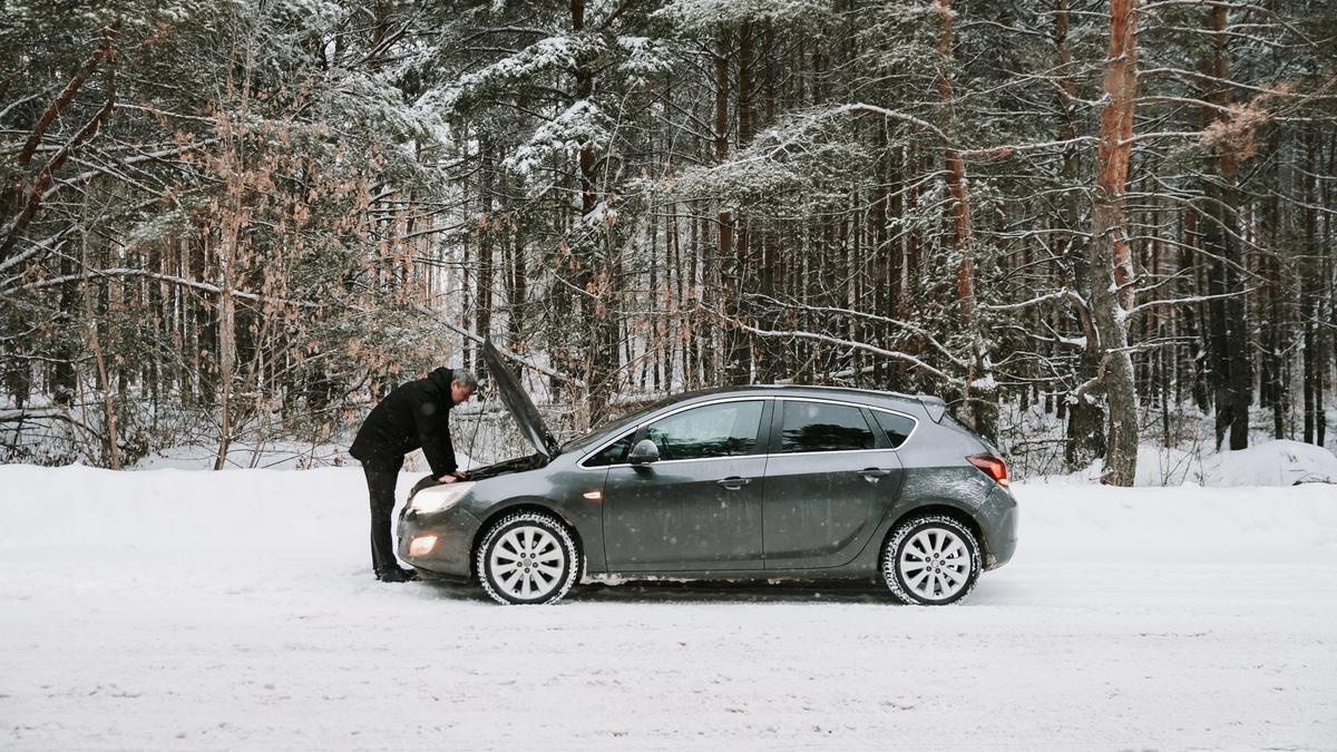 Person som undersøker motoren til en grå bil som ikke starter på en snødekt vei.