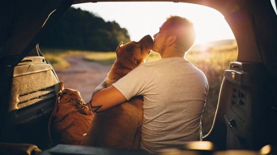 Mies istuu auton takapenkillä auringonlaskun aikaan ja halaa ruskeaa koiraa, joka nuolee hänen kasvojaan.