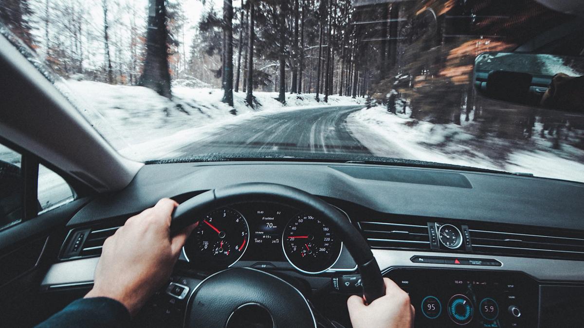 Näkymä auton sisältä, jossa kuljettaja ajaa lumisella, metsän reunustamalla tiellä, kädet ohjauspyörällä ja kojelauta näkyvissä.