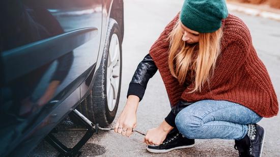 Vihreässä hatussa ja ruskeassa puserossa oleva henkilö nostaa autoa tunkilla.