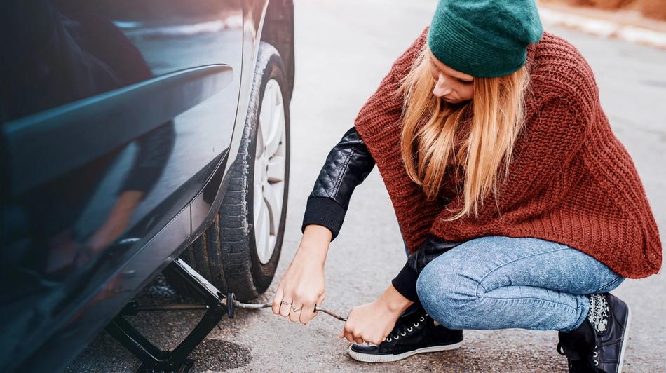 Vihreässä hatussa ja ruskeassa puserossa oleva henkilö nostaa autoa tunkilla.