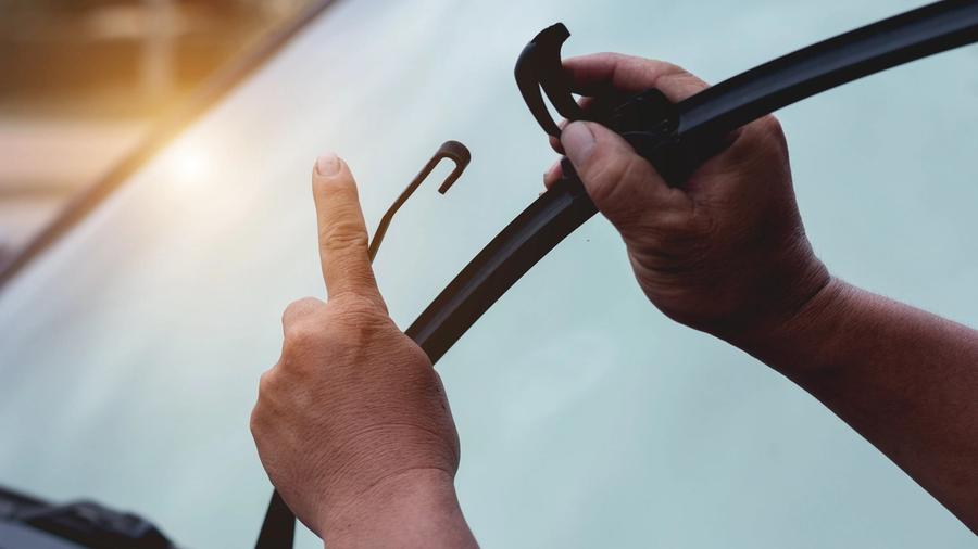 Henkilö vaihtaa koukkuvartista tuulilasinpyyhkijän sulkaa autossa, toisella kädellä pitäen sulaa ja toisella kädellä osoittaa koukkukiinnitystä.