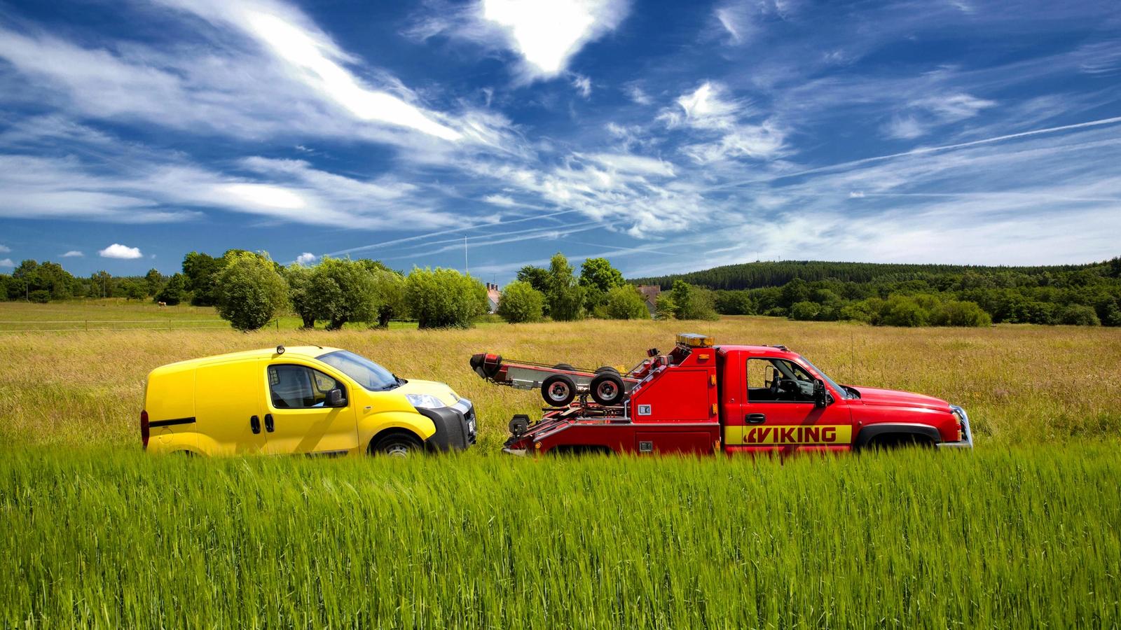 A red tow truck with the "Viking" logo is towing a small yellow van on a grassy field under a cloudy sky.