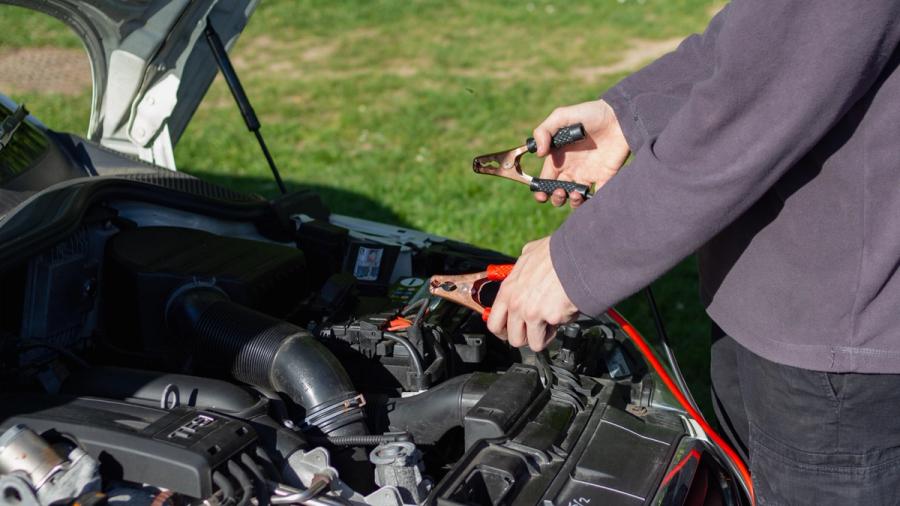 A person in front of the engine compartment of a car, holding the starter cables.
