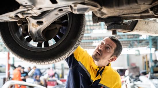 A mechanic in a yellow and blue uniform checks the bottom of a car that has been lifted up in the hall.