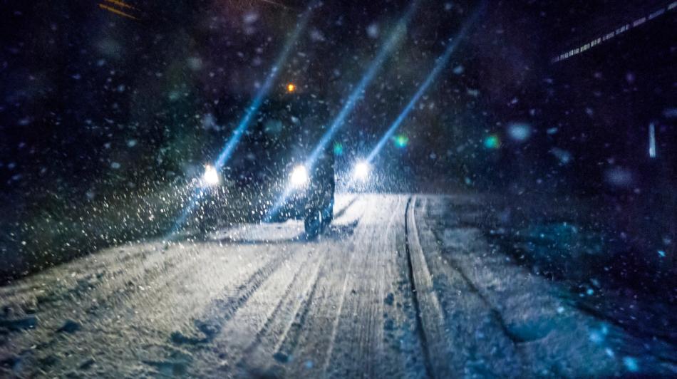 Auto ajaa yöllä lumipeitteisellä tiellä ajovalot päällä lumisateessa.
