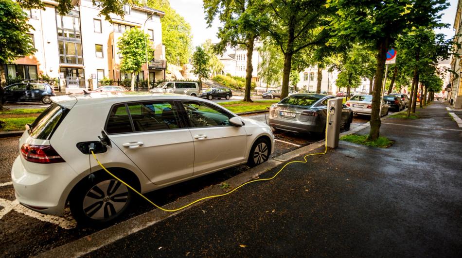 Valkoinen sähköauto on pysäköity kaupungin kadulle ja kytketty latauspisteeseen. Muita pysäköityjä autoja ja puita reunustaa märkää tietä.