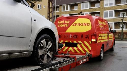 A Viking Assistance tow truck is towing a silver car with a flat tire in a residential area.