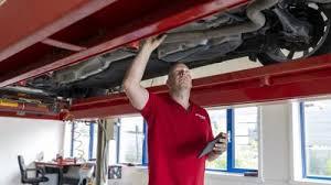 En mekaniker i röd skjorta inspekterar undersidan av en bil som lyfts upp på en hiss i ett garage, med en surfplatta i ena handen.