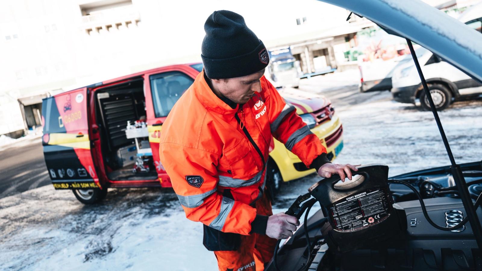 Oranssiin Viking työasuun pukeutunut henkilö liittää apukäynnistintä auton akkuun parkkialueella.