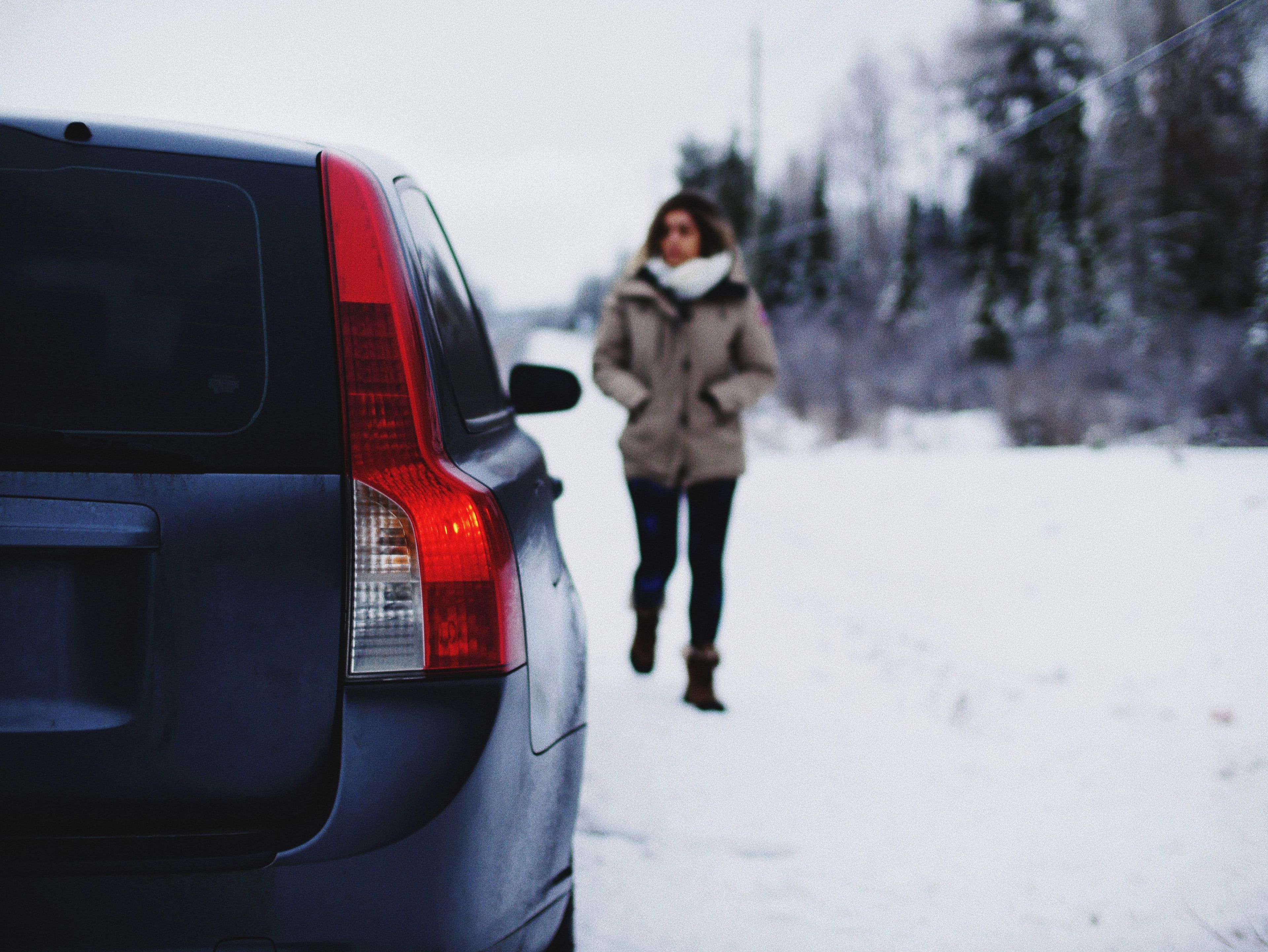Talvitakkiin pukeutunut henkilö kävelee lumisella tiellä lähellä pysäköityä tummanväristä autoa. Taustalla näkyy puita ja sähkölinjoja.