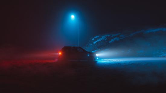 Auto, jonka ajovalot ovat päällä, on pysäköity yöllä sinisen katuvalojen alle, sumun ja lumipölyisen maiseman ympäröimänä.