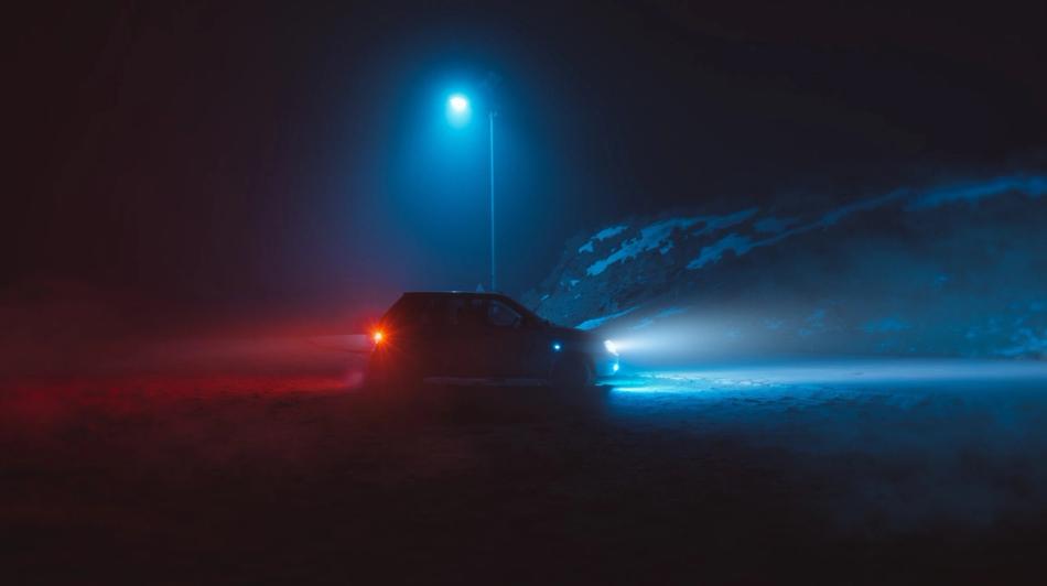 Auto, jonka ajovalot ovat päällä, on pysäköity yöllä sinisen katuvalojen alle, sumun ja lumipölyisen maiseman ympäröimänä.
