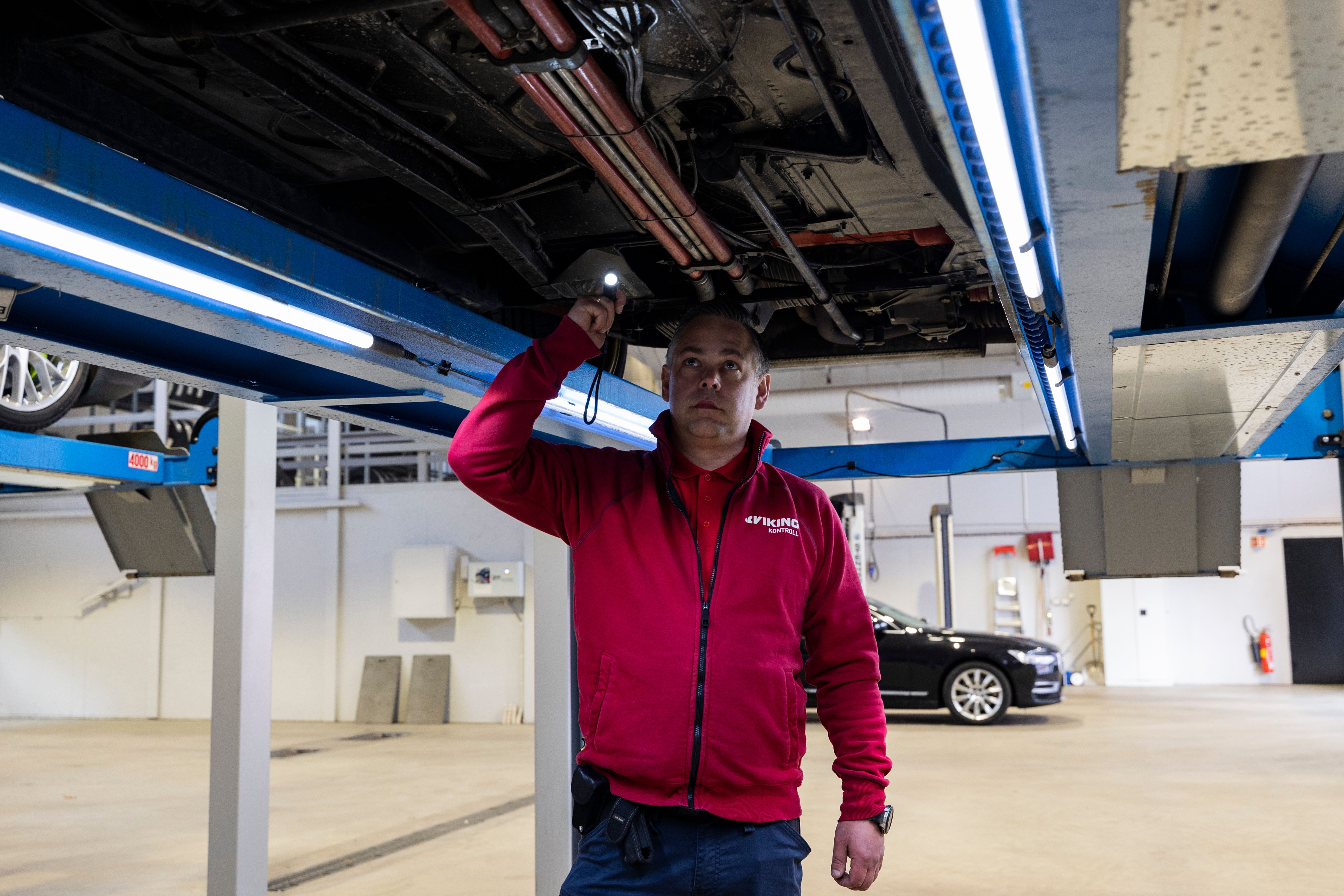 En mekaniker i röd skjorta inspekterar undersidan av en bil som lyfts upp på en hiss i ett garage, med en surfplatta i ena handen.