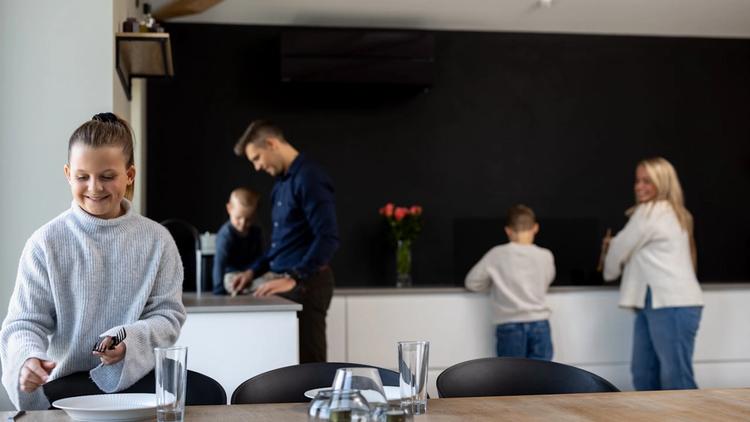 En familie på fire er på kjøkkenet. En jente dekker et bord, en mann og en gutt lager mat, og en kvinne står ved disken. Kjøkkenet har et trebord, svarte stoler og en vase med blomster.
