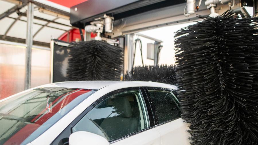 Valkoinen auto käy läpi automaattisen autopesun, jossa suuret pyörivät harjat ovat kosketuksissa ajoneuvon pintaan.