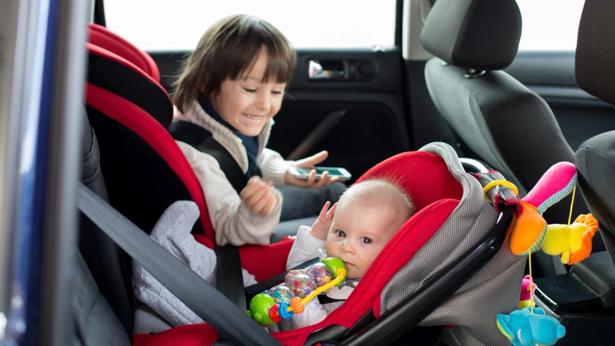 Pieni lapsi hymyilee auton takapenkillä kasvot menosuuntaan asennetussa turvaistuimessa vierellään vauva, joka on selkä menosuuntaan asennetussa turvakaukalossa.