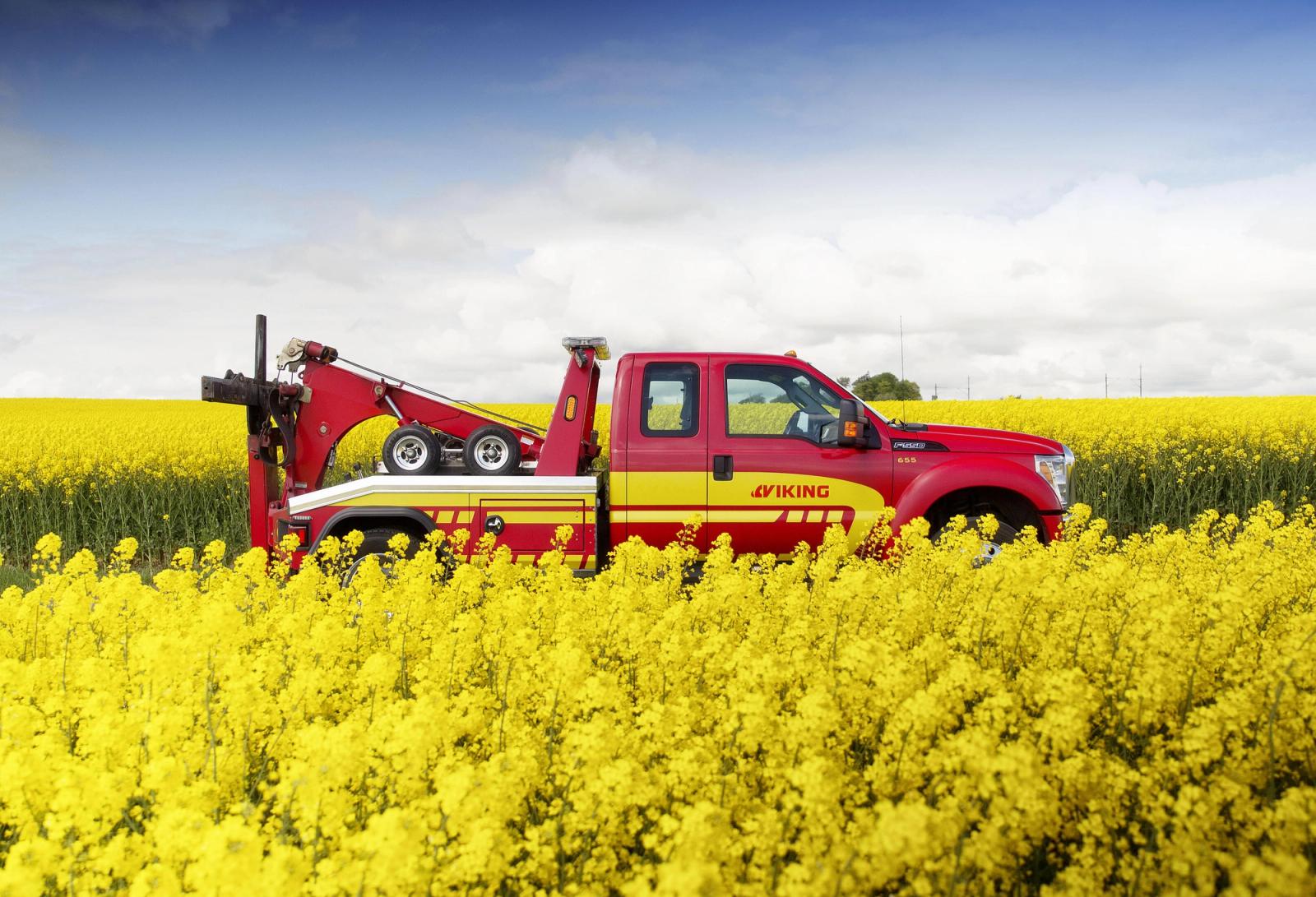 En röd bärgningsbil står parkerad på ett fält med klargula blommor under en blå himmel med spridda moln.