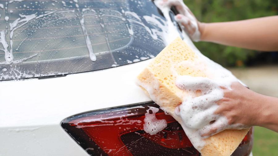 En person som bruker en såpesvamp for å vaske bakvinduet og baklyset på en hvit bil som forebyggende tiltak mot rust.