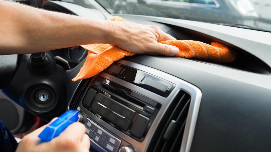 Henkilö puhdistaa auton kojelautaa oranssilla kankaalla ja suihkepullolla.