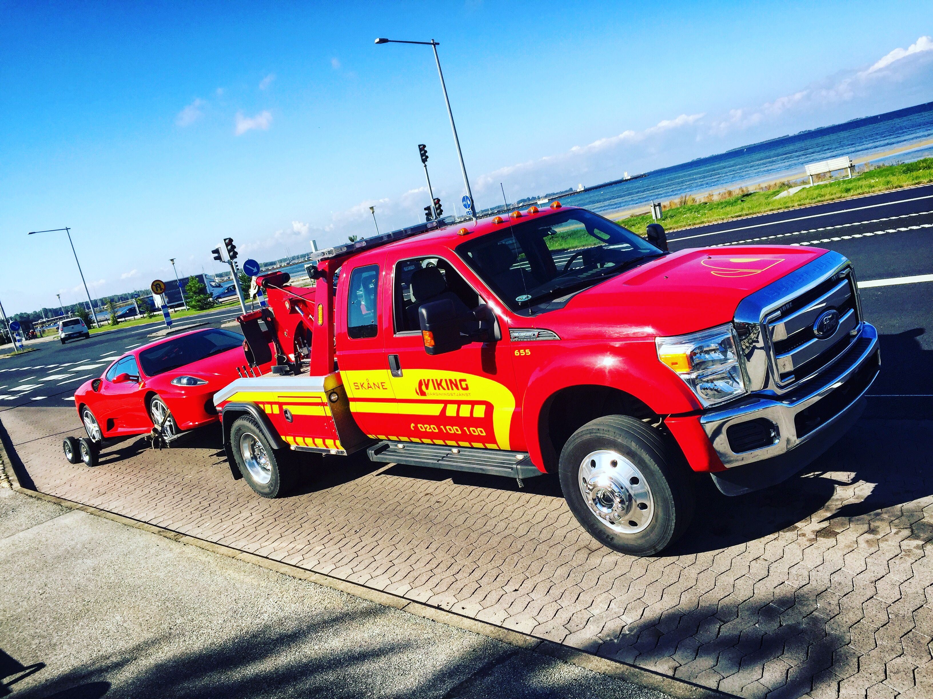 Punainen hinausauto hinaa punaista autoa tiellä lähellä rantaviivaa kirkkaana, aurinkoisena päivänä.