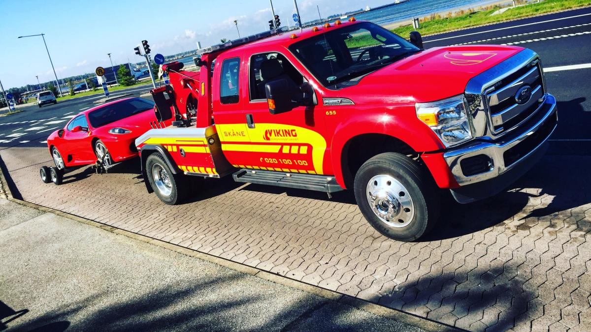 Punainen hinausauto hinaa punaista autoa tiellä lähellä rantaviivaa kirkkaana, aurinkoisena päivänä.