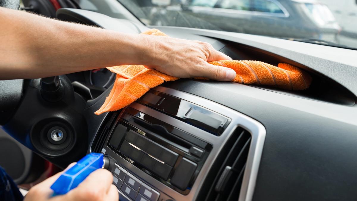 Henkilö pyyhkii auton kojelautaa oranssilla kankaalla pitäen sinistä suihkepulloa.