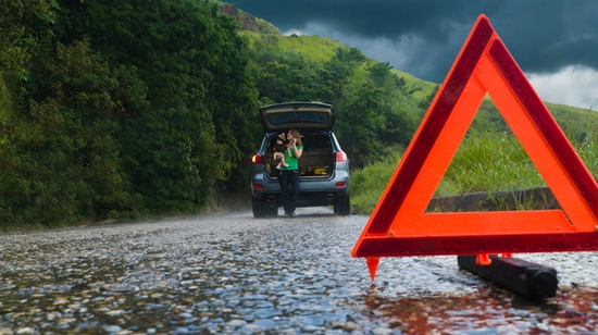 Henkilö seisoo avoimen auton tavaratilan vieressä märän tien varrella, ja etualalla on maahan asetettu punainen varoituskolmio.