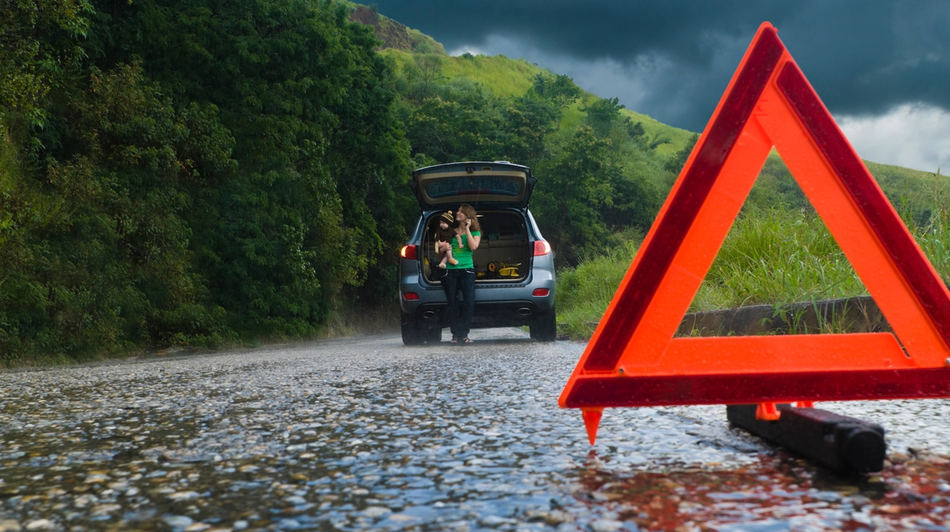 Henkilö seisoo avoimen auton tavaratilan vieressä märän tien varrella, ja etualalla on maahan asetettu punainen varoituskolmio.