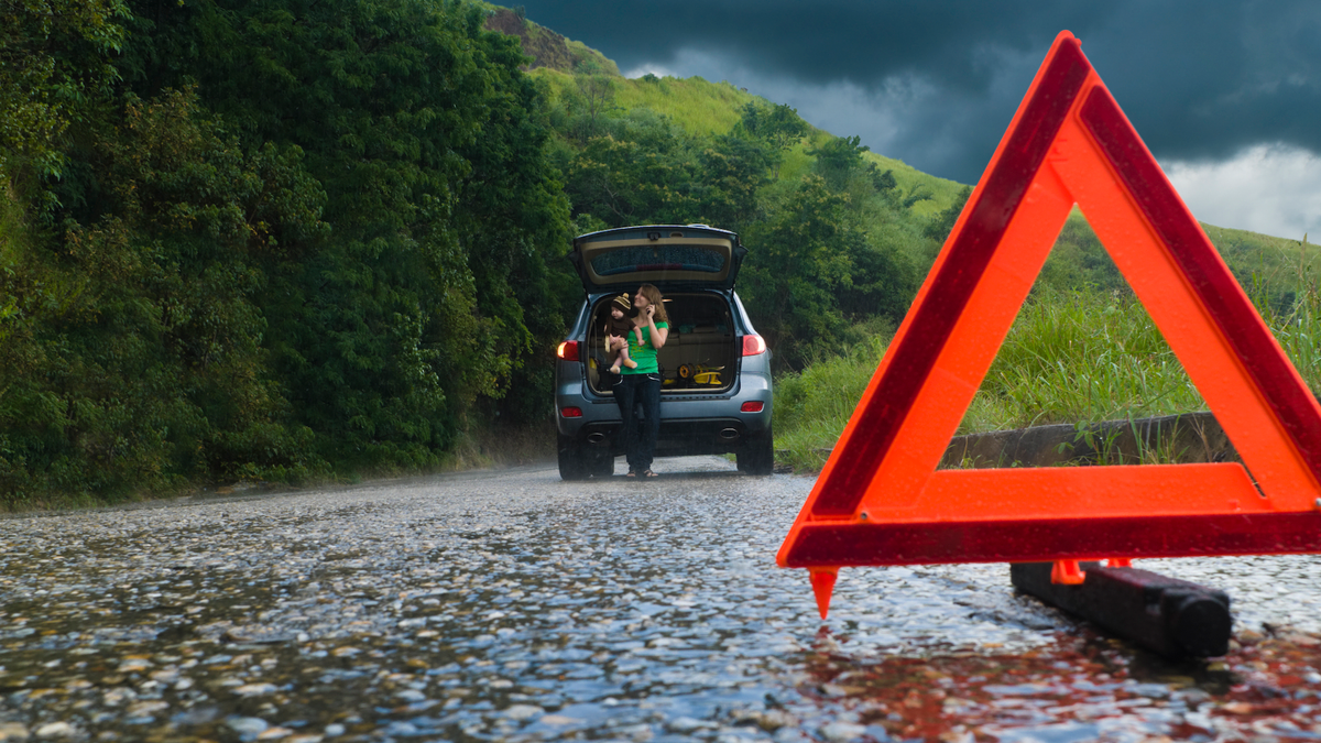 Henkilö seisoo avoimen auton tavaratilan vieressä märän tien varrella, ja etualalla on maahan asetettu punainen varoituskolmio.
