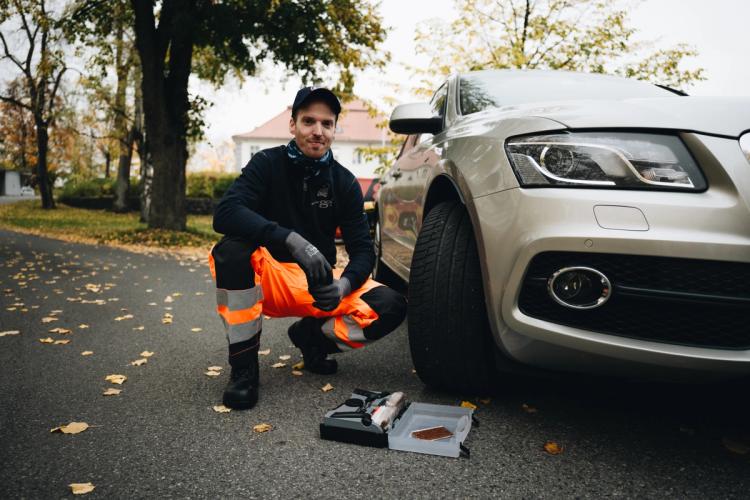 Mies turvavarusteissa polvistumassa auton vieressä puiden reunustamalla kadulla, kädessään työkalu avoimesta työkalulaatikosta eturenkaan lähellä.