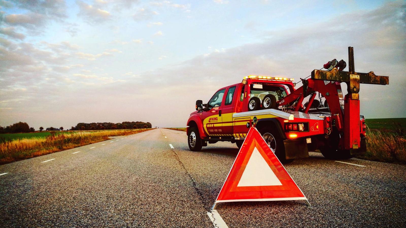 A red Viking tow truck parked on a rural road with a warning triangle in the foreground, surrounded by open fields under a cloudy sky.