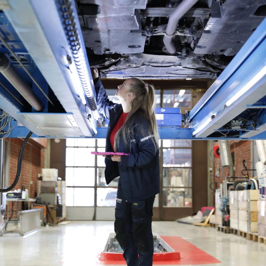 Mekaanikko tarkastaa autokorjaamossa nostimella olevan auton alapuolta vaaleanpunainen leikepöytä kädessään.