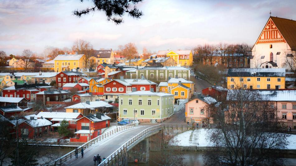 Porvoon jokirannan maisemaa, jossa on värikkäitä puutaloja, joen ylittävä silta ja suuri kirkko talvipäivänä.