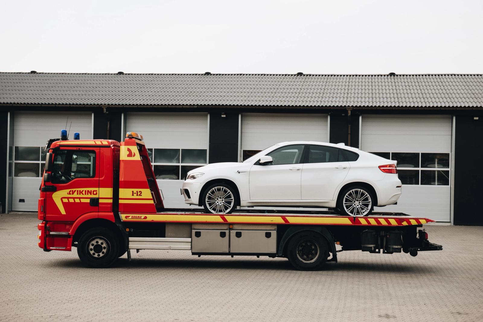 Valkoinen auto Vikingin hinausauton kyydissä, joka on pysäköity autotallin ovilla varustetun rakennuksen eteen.