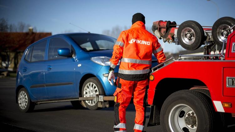 A tow truck driver in an orange Viking uniform is loading a blue car onto a tow truck.