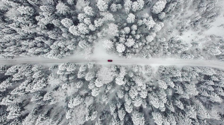 Ilmakuva lumen peittämästä metsästä, jonka läpi kulkevalla lumisella tiellä ajaa punainen auto.
