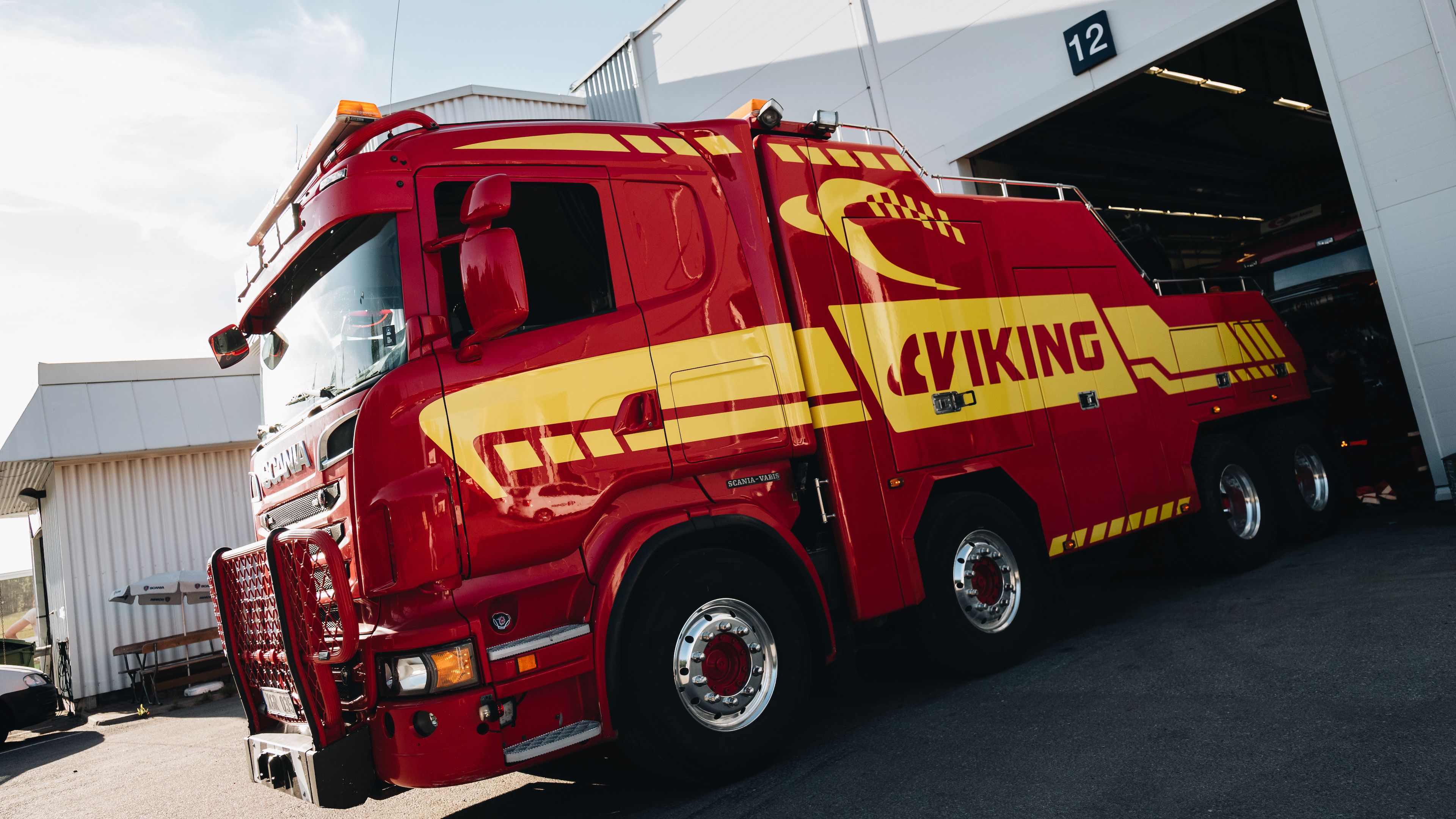 Suuri punainen ja keltainen hinausauto, jossa lukee "VIKING", on pysäköity autotallin ulkopuolelle, jossa on numero 12.