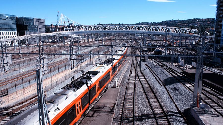 Et rødt og hvitt tog kjører gjennom en jernbanestasjon med flere spor og luftledninger, omgitt av urbane og industrielle bygninger.