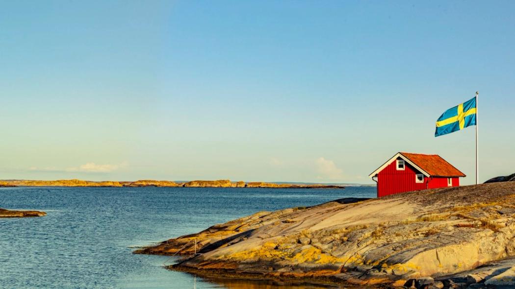 Rött hus med vita knutar och en flaggstång med en svensk flagga hissad, på en ö i skärgården.