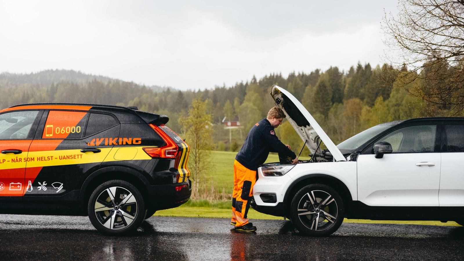 En Viking bilberger i synlig uniform inspiserer motoren til en hvit bil på en regnværsdag, med et servicekjøretøy parkert i nærheten.