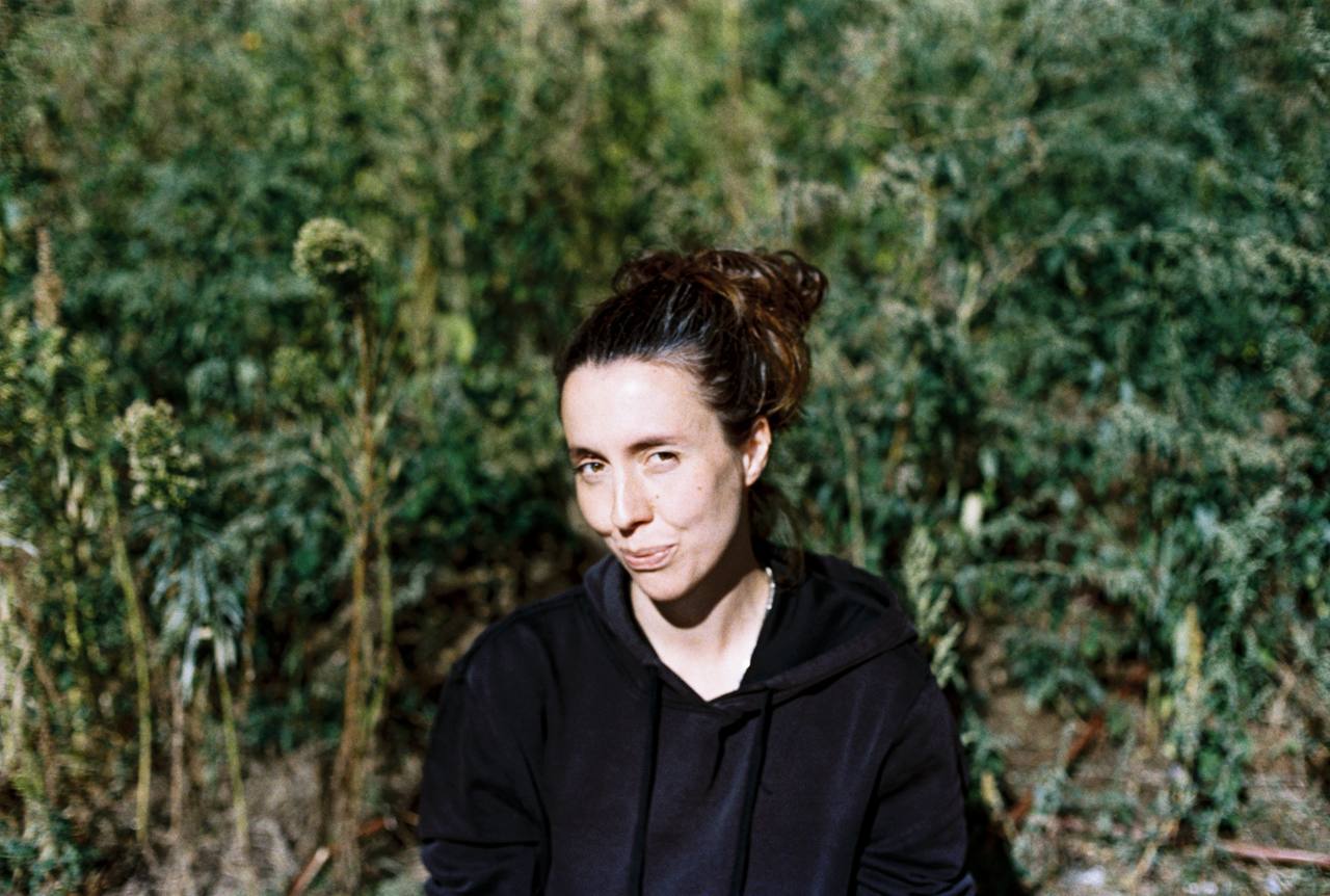 A person with brown hair tied up, wearing a black hoodie, sits outdoors in front of green, bushy plants, looking at the camera.