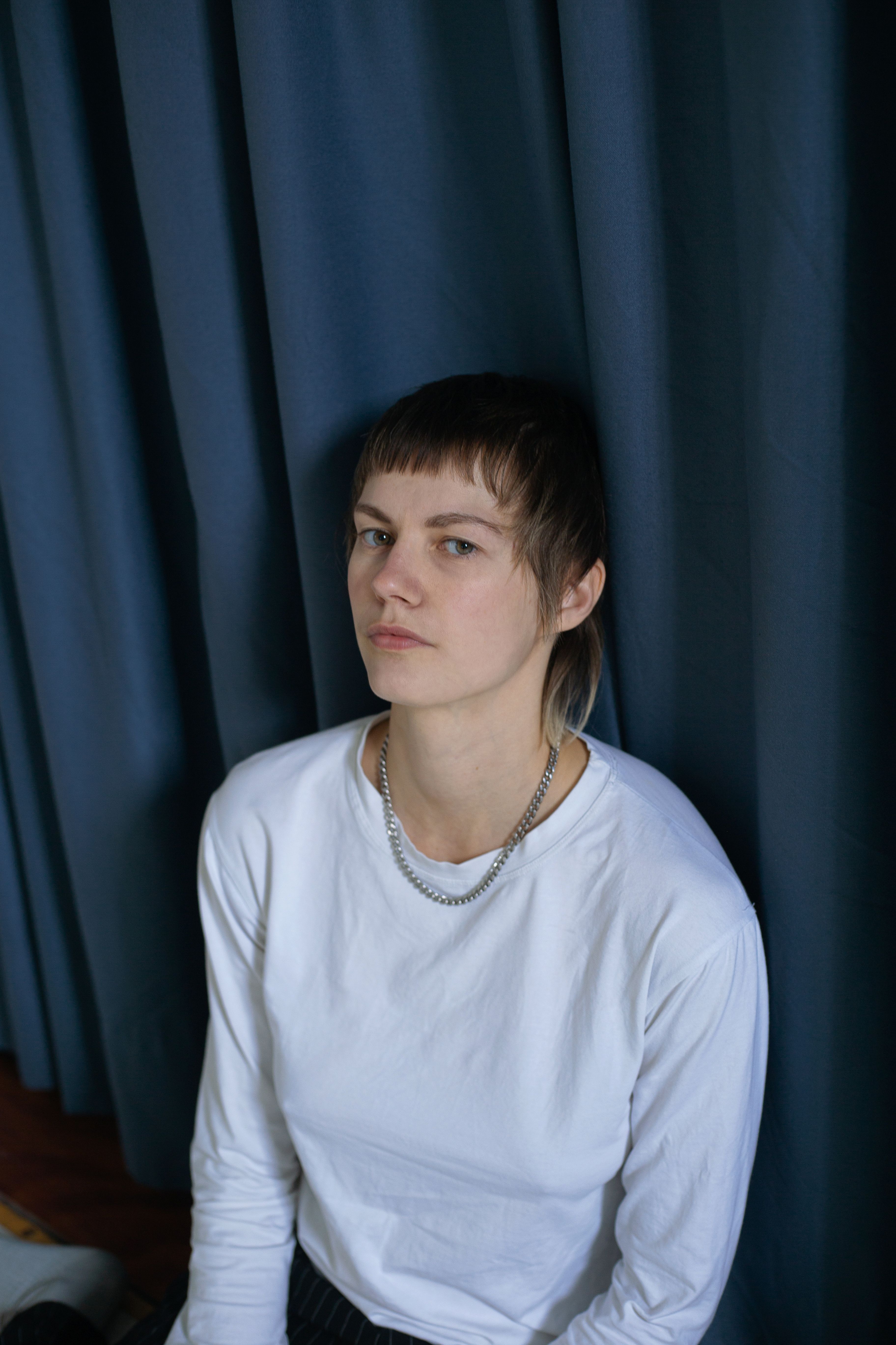 Person with white shirt, silver chain, bangs, leaning against a blue curtain