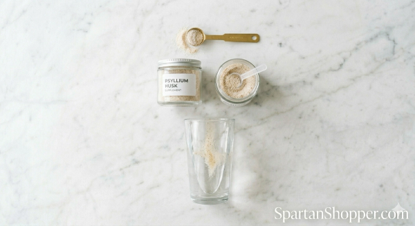 Measuring spoon, psyllium husk powder jar, and glass of water on a white marble kitchen counter showing how to mix psyllium husk